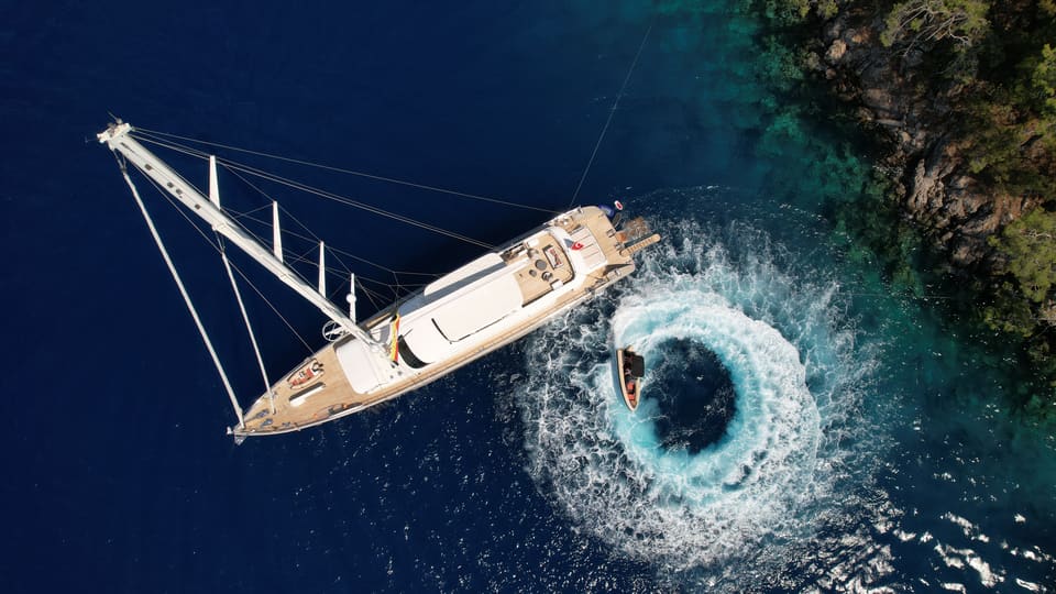 2023 Custom Laminated Motorsailer 41M – aerial view of the sailing yacht at anchor near a rocky shore, tender circling in clear blue water