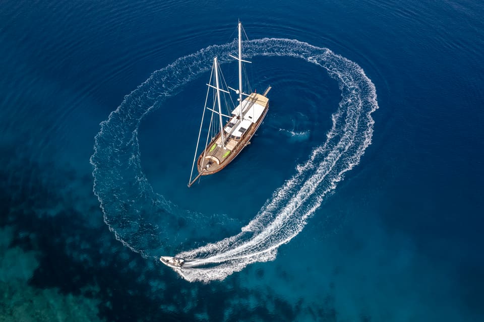 2009 Custom Ketch 31M – aerial view of the wooden ketch at anchor on deep blue water, with a tender circling and leaving a white wake