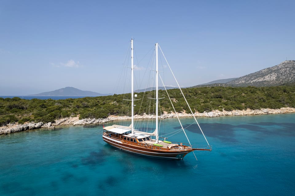 2009 Custom Ketch 31M – aerial view of wooden twin-mast gulet at anchor in turquoise water off a green coastline