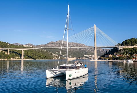 2016 Lagoon 400 S2 – sakin suda motorla seyreden katamaran, arka planda askılı köprü ve tepeler