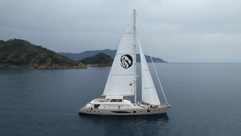 2023 Custom Laminated Motorsailer 41M – aerial view under full sail with mainsail and headsail on calm water near green hills